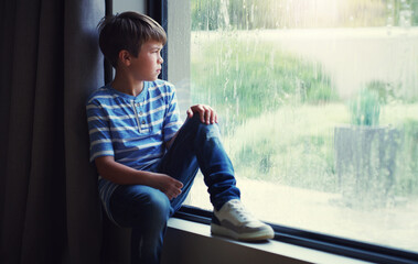 Little boy, home and watching the rain by window of natural water drops and thinking in relax indoors. Child relaxing by windowsill looking out glass at storm in lonely or sad for weather at house