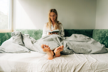A woman is reading while sitting in bed. Blurred portrait of a woman, focus on the feet.
