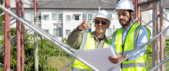 Two young architect discussion or contractor man while looking blueprint building structure together for survey on construction site, engineer planning project, industry and construction concept.
