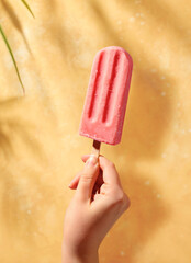 Woman hand holding pink fruit popsicle on sunny yellow background