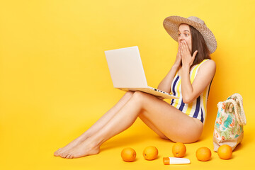 Shocked surprised woman wearing striped swimming suit and hat isolated on yellow background working...