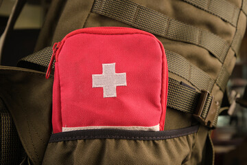 A red first aid kit with a white cross in the pocket of a tactical military olive backpack with a Molle system - obrazy, fototapety, plakaty
