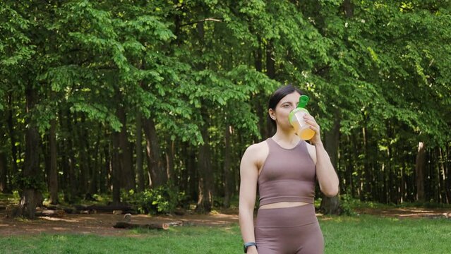 Athletic woman drinking sport nutrition with bcaa amino acids and vitamins from shaker, outdoors in summer park after morning workout, slow motion orbit motion, healthy lifestyle concept