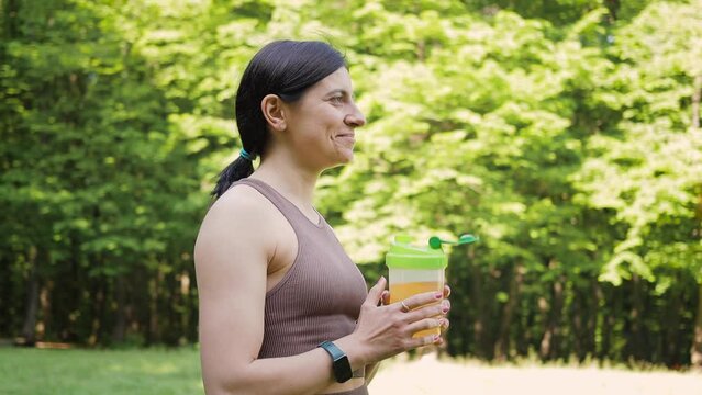 Athletic woman drinking sport nutrition with bcaa amino acids and vitamins from shaker, outdoors in summer park after morning workout, slow motion orbit motion, healthy lifestyle concept