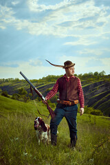 Portrait with man, vintage hunter wearing retro clothes with hat with feather holding gun and standing with dog English springer spaniel over nature landscape background
