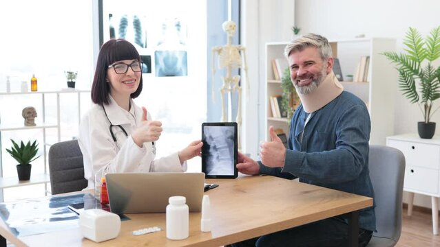 Mature bearded man in C collar talking to female physician in white coat pointing at tablet with spine scan on screen showing thumb up