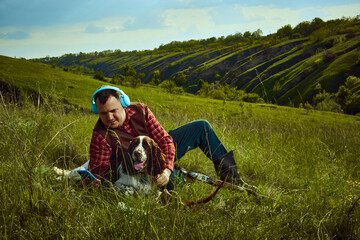 Portrait with man, hunter sitting on grass with his dog English springer spaniel listening to music and resting after hunting on nature landscape background