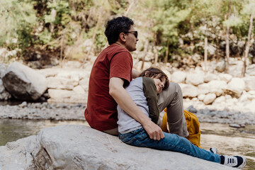 School boy and his dad with yellow backpack sits on a riverside rock in the canyon with mountain cliffs in the background. Kid child and father taking a rest on a boulder near mountain river.