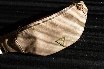 Glamorous women's waist bag in gold color on a black shiny background. Fashionable handbag with gold chain.