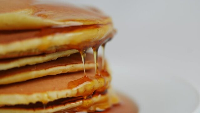 Pile of pancakes on the left with golden honey drops flowing down in slow motion. Delicious sweet brunch dessert at white background close-up