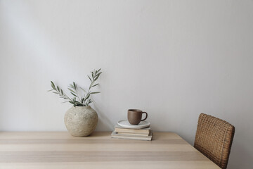 Neutral Mediterranean home design. Vintage vase with olive tree branches, cup of coffee. Books on wooden table, rattan chair. Living room still life. Empty wall copy space. Modern interior, no people. - obrazy, fototapety, plakaty
