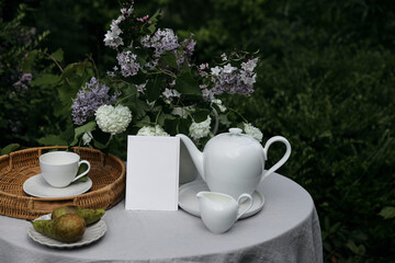 Closeup of blank greeting card, birthday wedding invitation mockup. Tea party in green garden. Teapot, milk pitcher, pear fruit on table with linen table cloth. Cup of coffee. Lilacs, viburnum flowers