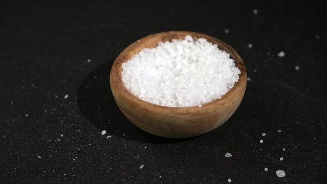 A girl dials salt into a wooden spoon from a bowl on a black background. Coarse sea salt.