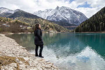 Fototapeta na wymiar Woman in black clothes standing on lakeside of Auronzo Lake, in Auronzo di Cadore village; Snow peaked mountains on background; Belluno, Dolomites, Italy
