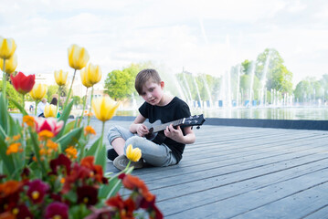 a little boy sits by the fountain, plays the black ukulele on a sunny day in the park	