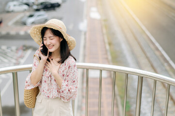 young asian woman traveler with weaving basket using mobile phone and standing on overpass with railway background. Journey trip lifestyle, world travel explorer or Asia summer tourism concept.