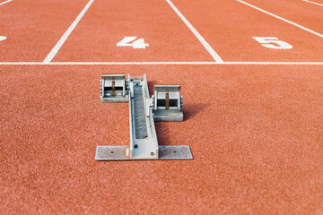 Red textured running track surface with a starting block in front of the start line number four, ground level close up shot.