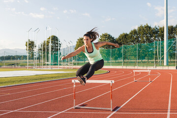 Young overweight woman running and jumping over hurdles on an athletics track on a sunny summer day