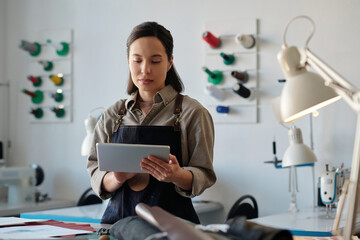 Young serious female designer looking at tablet screen by workplace while scrolling through new online models of fashionable apparel