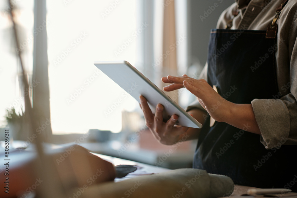Poster Hands of young female tailor or clothes designer using tablet in workshop while standing in front of camera and looking through online orders
