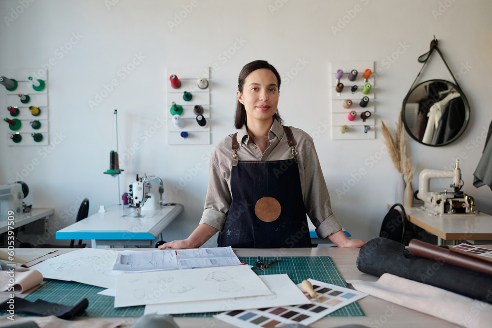 Poster young pretty leatherworker in apron looking at camera while standing by workplace with sketches, fab