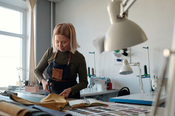 Professional female designer of clothes cutting piece of leather by table with open notebook with...
