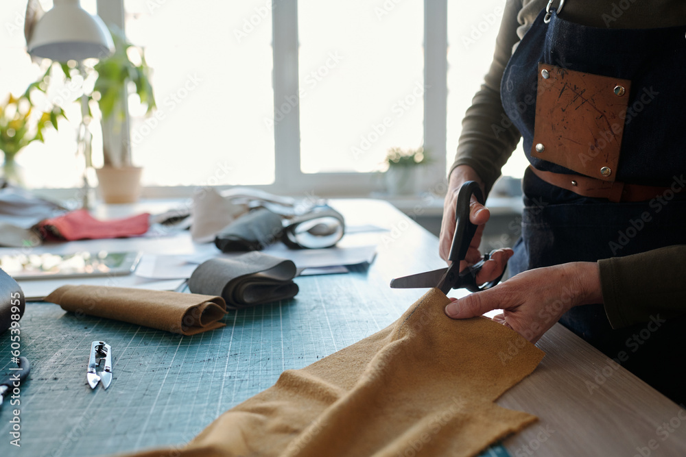 Poster close-up of female leatherworker in apron cutting leather or suede while standing by table in front 