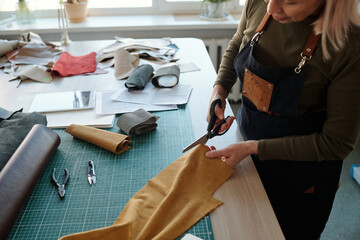 Cropped shot of mature female leatherworker cutting piece of suede or leather with scissors while working over new creative apparel - obrazy, fototapety, plakaty