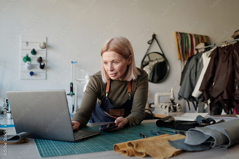 Poster blond mature owner of leatherworking shop looking at laptop screen while standing by workplace and s