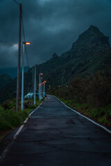 Moody picture of a street going uphill, dark blue sky and orange streetlights and big mountain in the background