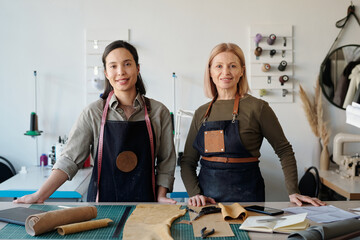 Happy young and mature craftswomen in workwear looking at camera while standing by workplace with pieces of textile in workshop