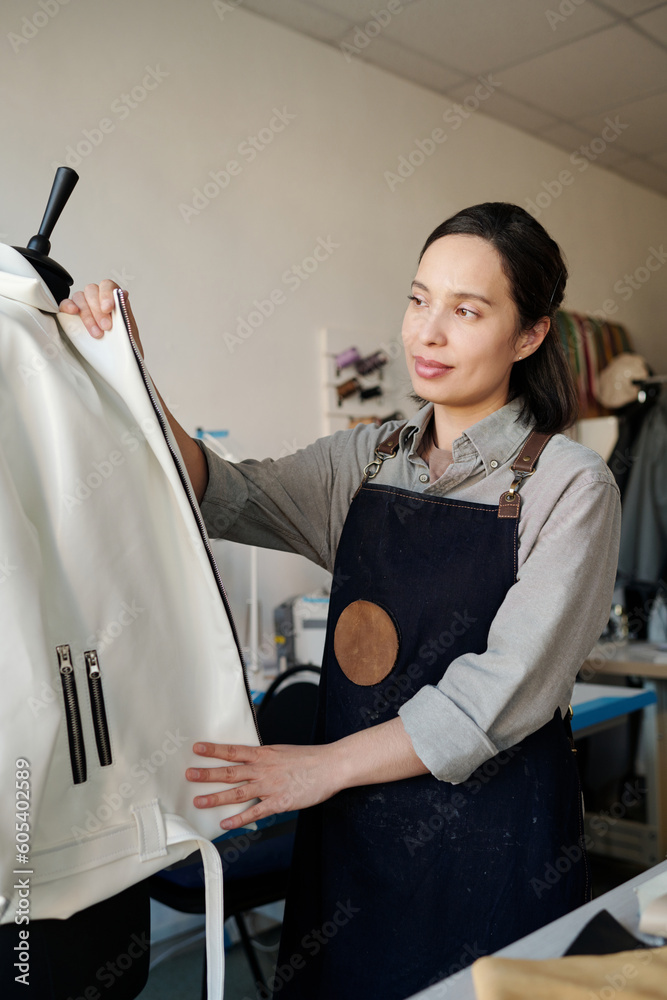 Poster young pretty tailor standing in front of mannequin with new white leather jacket while working over 