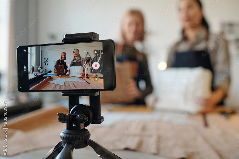 Poster Two creative craftswomen on screen of smartphone standing on tripod in front of camera during masterclass on creating leatherwear