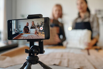 Two creative craftswomen on screen of smartphone standing on tripod in front of camera during masterclass on creating leatherwear