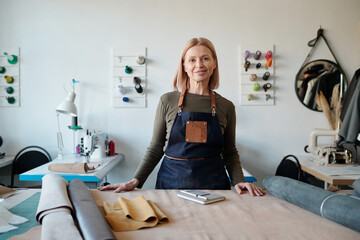 Smiling mature craftswoman in apron looking at camera in workshop or studio while standing by table...