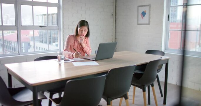 Focused asian businesswoman using laptop and taking notes, at table in meeting room, in slow motion
