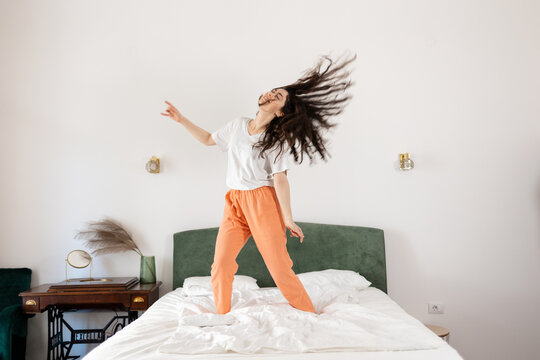 Happy young Caucasian woman dancing on bed in bedroom. Dark hair fly away. Freedom and relax at home.