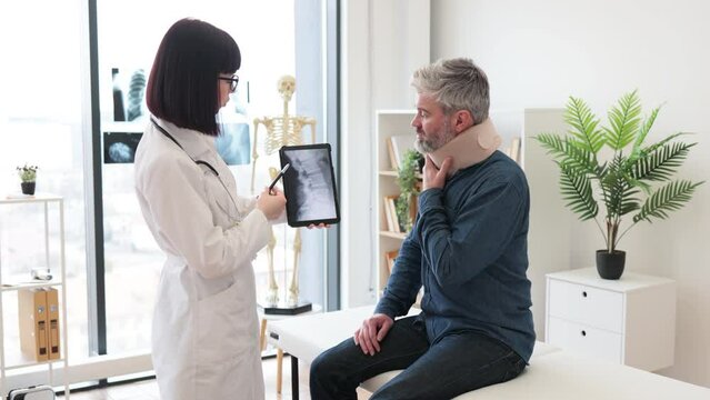 Caucasian lady holding tablet with CT scan image on screen while talking to male in neck brace sitting on exam couch. Young orthopedist analyzing thoracic vertebrae on digital report in clinic.