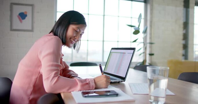 Happy asian businesswoman using laptop and making notes, at table in meeting room, in slow motion