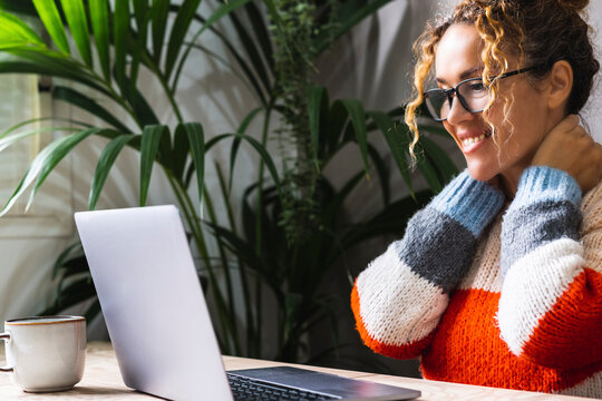 Happy Woman Smile And Look At Laptop Screen Touching Her Neck. Modern People Using Notebook Computer On The Table. Portrait Of Lady Wearing Eyeglasses. Concept Of Online Activity And Enjoyment Lady