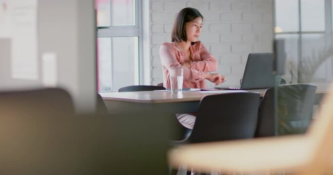 Focused asian businesswoman looking at laptop at table in empty meeting room, in slow motion