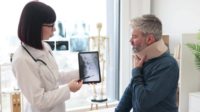 Caucasian lady holding tablet with CT scan image on screen while talking to male in neck brace sitting on exam couch. Young orthopedist analyzing thoracic vertebrae on digital report in clinic.
