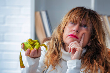 pensive middle aged woman looking at measuring tape, diet and health concept