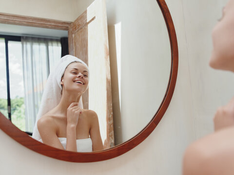 A beauty woman stands in front of a mirror after a shower in a towel on her head looks at her reflection and does a facial massage applies a day cream, beauty facial skin care smile, anti acne