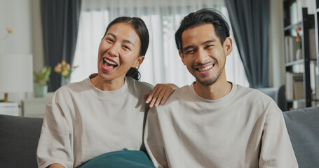 Happy young Asian couple sit on couch smiling looking at camera talk video call with friend and family at home on holiday. Smile young husband and wife webcam conversation using internet connection.