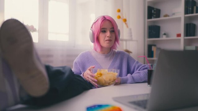 Young woman with pink hair eating potato chips and watching a movie at home