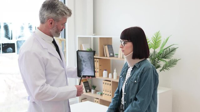 Caucasian woman in neck brace looking at x-ray image on digital device held by bearded therapist in lab coat. Medical specialist explaining test results to patient with walking cane in exam room.