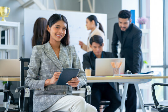 Portrait of pretty business woman sit on chair also hold tablet and look at camera with smiling in office and other co-worker work in the back.