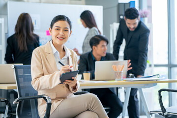 Portrait of business woman sit on chair also hold tablet and look at camera with smiling in office and other co-worker work in the back.