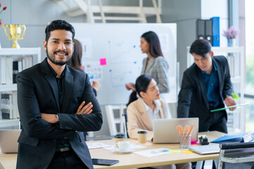 Portrait of Asian business man with beard stand with arm crossed also smiling look at camera and stay in front of other co-worker work in the back in office.